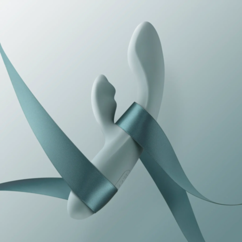 The Svakom-Chika G Spot Warming Rabbit in Turquoise Grey is elegantly designed with sleek, abstract curves that evoke the form of a clitoral stimulator. It features a smooth, flowing blue ribbon partially wrapping around it and is set against a gradient b