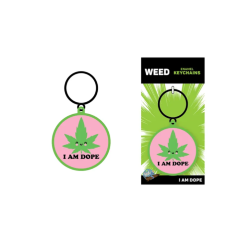 Keychain with a circular design and a pink center showcasing a cartoon green leaf with eyes, accompanied by the text "I AM DOPE." It is displayed in its packaging labeled "WEED Enamel Keychains," featuring a green, spiky background. The product is named W