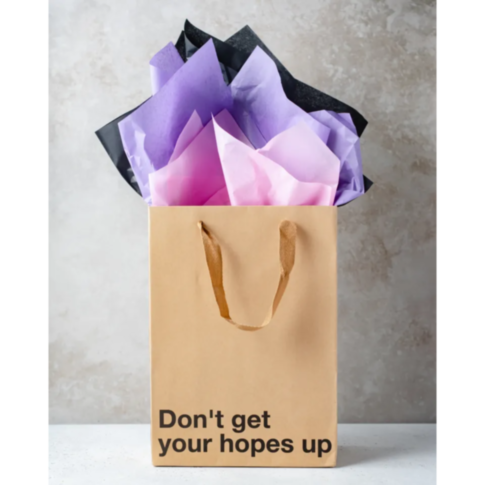 Introducing the Wood Rocket-Don't Get Your Hopes Up Bag, a snarky gift bag featuring sturdy handles and filled with crumpled pink, purple, and black tissue paper. Its design includes the phrase 
