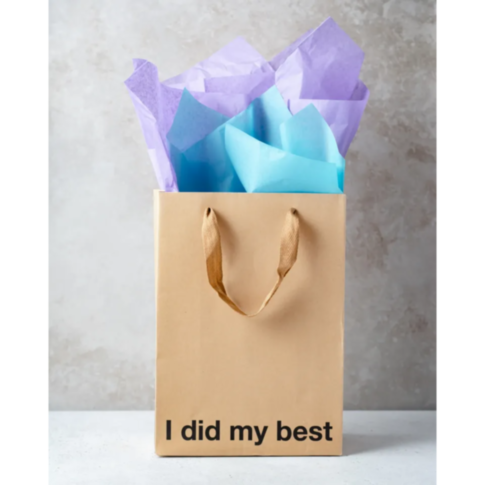A sarcastic gift bag from Wood Rocket, called the "I Did My Best Bag," features two handles and holds purple and blue tissue paper. It rests on a white surface against a textured gray background with the phrase "I did my best" emblazoned on the front.