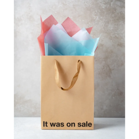 The Wood Rocket-It Was On Sale Bag, a brown paper gift bag with handles, is filled with vibrant red and blue tissue paper and stands confidently against a gray textured background. The playful and snarky message "It Was On Sale" is boldly printed at the b