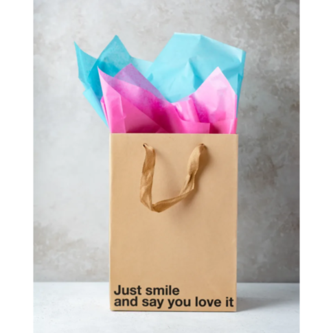 The Wood Rocket-Just Smile Bag, a beige gift bag featuring brown handles, is whimsically filled with blue and pink tissue paper. The humorous text "Just smile and say you love it" is printed on the bag, making it ideal for gag gifts. It sits against a neu