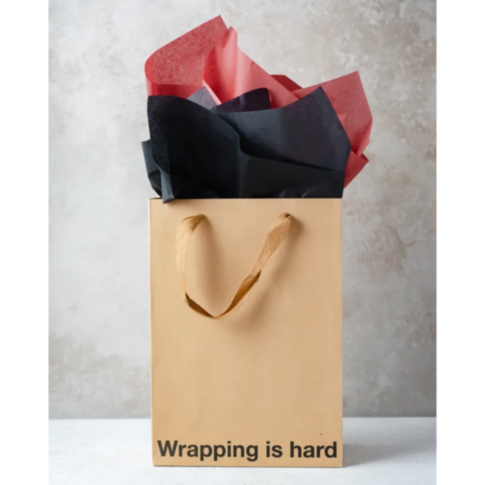 A Wood Rocket "Wrapping Is Hard" bag, featuring crumpled red and black tissue paper spilling out from the top, is set against a light textured background. The humorous text at the bottom of the image reads, "Wrapping is hard," perfectly encapsulating a bi
