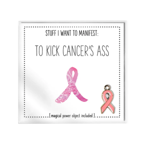 A square image features the bold text "STUFF I WANT TO MANIFEST: TO KICK CANCER'S ASS." Beneath it, an illustration of a pink ribbon and an accompanying charm symbolize hope. The caption states [Magical Power Object included]. This empowering "To Kick Can
