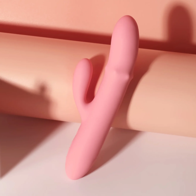 A Svakom-Mora Neo Thrusting Beaded Ring Rabbit in Peach Pink, featuring a dual-pronged design, is positioned upright against a rolled peach-colored background, casting a gentle shadow. The ambient light enhances the warm tones in this minimalist compositi