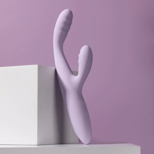 The Svakom-CiCi 2+ Heating Rabbit-Lilac, a beginner-friendly vibrator designed for luxurious G-spot stimulation, leans elegantly against a white cube. It showcases a curved design with textured grips and buttons, all set against a soft lilac background fo