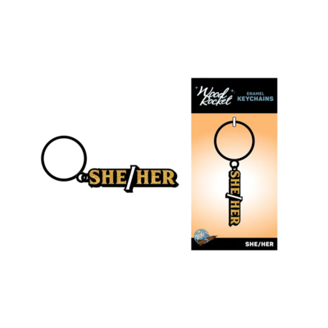 The Wood Rocket-She/Her Keychain showcases the words SHE/HER in striking black and gold enamel, enhanced by a stunning glitter accent. The packaging, visible on the right and labeled Wood Rocket Enamel Keychains, offers a close-up view of this elegant des