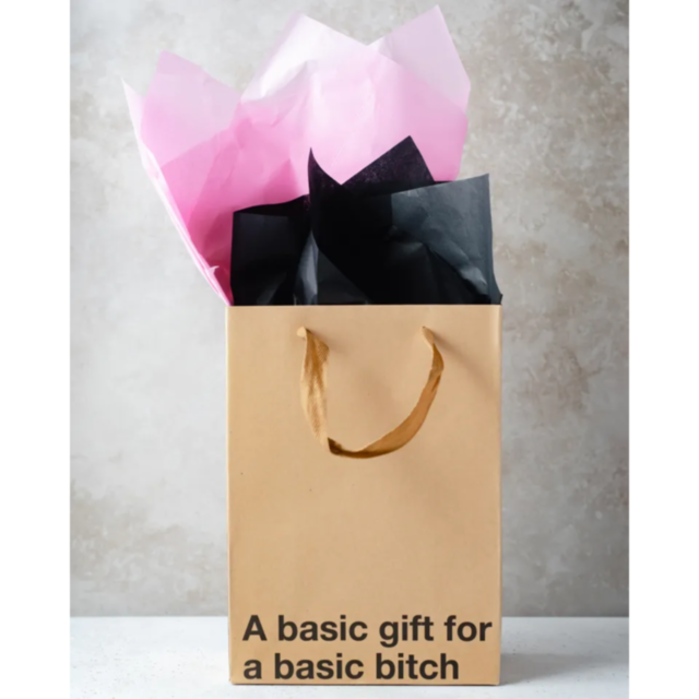 The "Wood Rocket-A Basic Gift Bag," showcasing pink and black tissue paper, humorously displays the phrase "A basic gift for a basic bitch." Ideal for a White Elephant exchange, it rests against a light gray stone background.