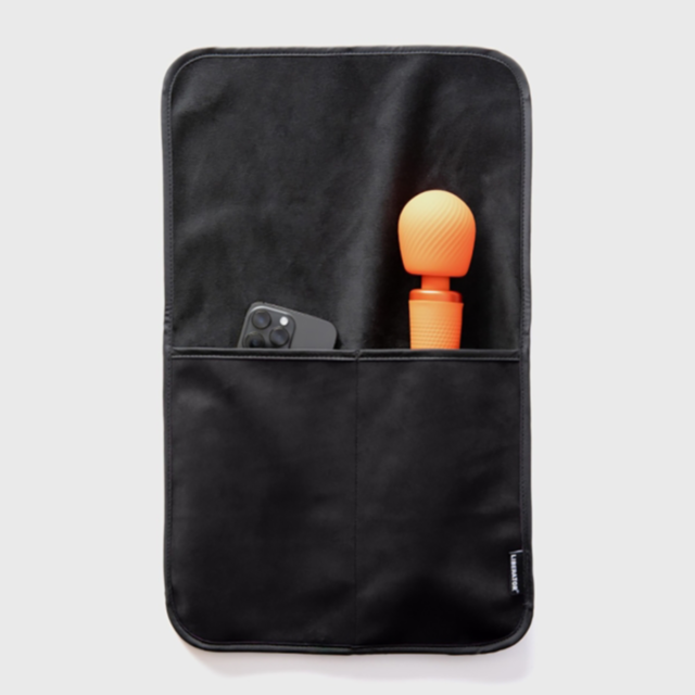 The Liberator Fascinator Bed Caddy in black microvelvet features two pockets. One pocket securely holds a smartphone encased in dark armor, while the other accommodates an orange, textured item with a rounded top. The scene is set against a plain white ba